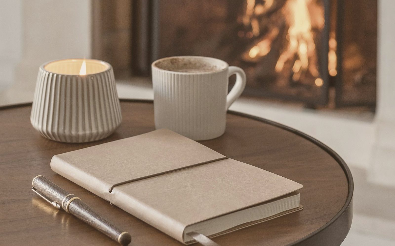 A notebook, pen, coffee cup and candle on a walnut table by a fireplace